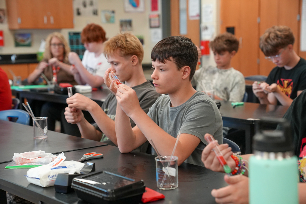 A student carefully pours clear liquid into a test tube containing a red strawberry mixture during a DNA extraction lab.