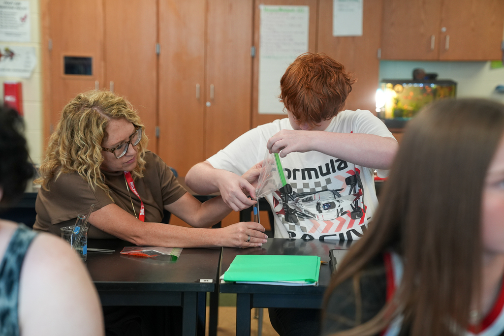 A student transfers liquid from a plastic bag into a test tube while an adult steadies the tube during a strawberry DNA extraction lab.