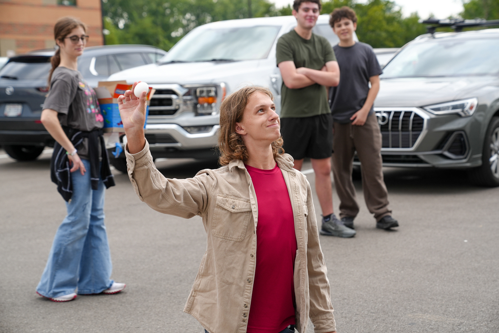 A student holds up a white egg and looks upward during the outdoor egg drop activity as classmates watch nearby.