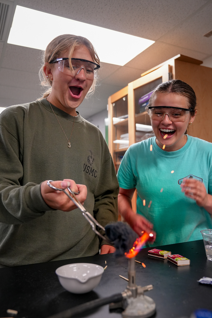 Two students wearing safety goggles react with excitement as one holds steel wool over a Bunsen burner with tongs, producing bright orange sparks.