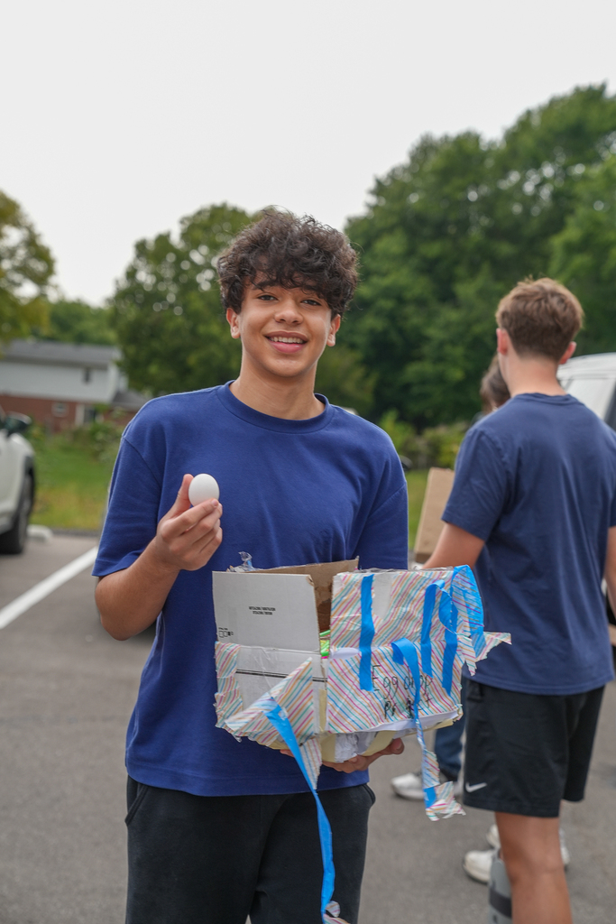 A smiling student displays a white egg alongside a homemade protective chamber made from cardboard, colorful paper and blue tape.