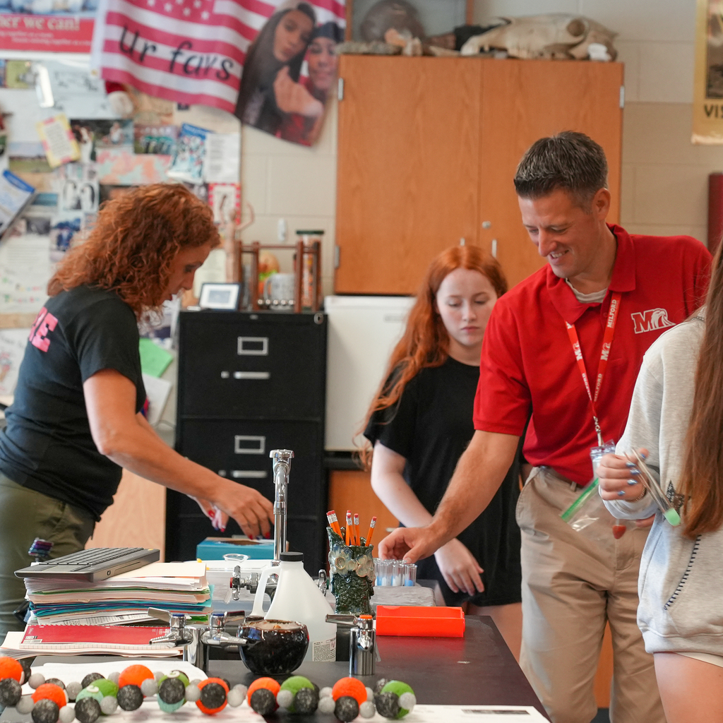 An adult wearing a red Milford polo smiles and reaches toward a test tube rack as students and another adult gather around a lab station during the strawberry DNA extraction activity.