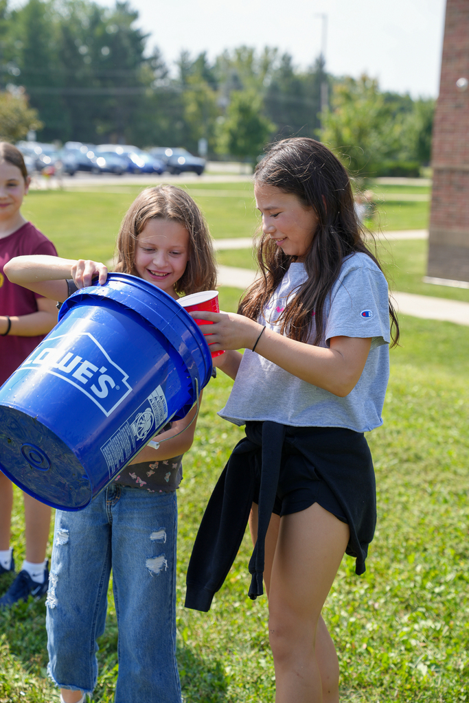 Two Seipelt students smile as they fill red cups with water from a large blue bucket.