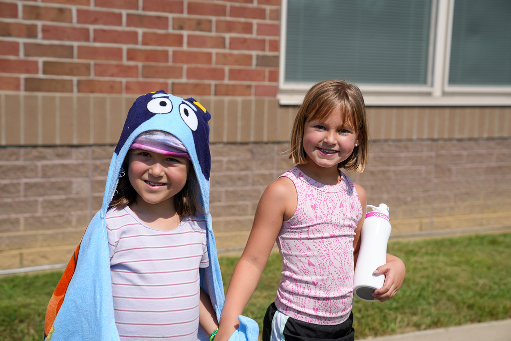 Two smiling Seipelt students hold hands outside, with one wearing a colorful character towel and the other carrying a water bottle.