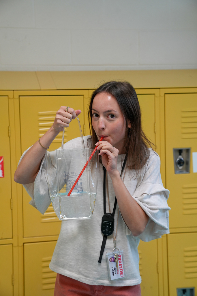 A staff member smiles while drinking through a straw from a clear plastic tote bag in front of yellow lockers.