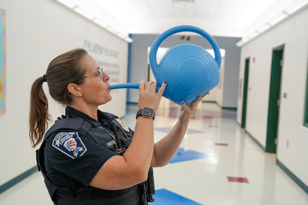 A Miami Township police officer playfully drinks from the spout of a large blue watering can in a school hallway.