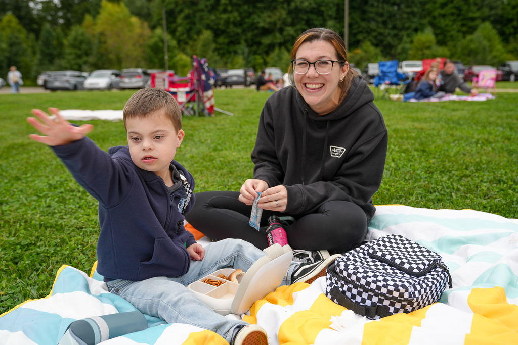 A student gestures while seated beside a smiling adult and an open lunch box on a blanket.