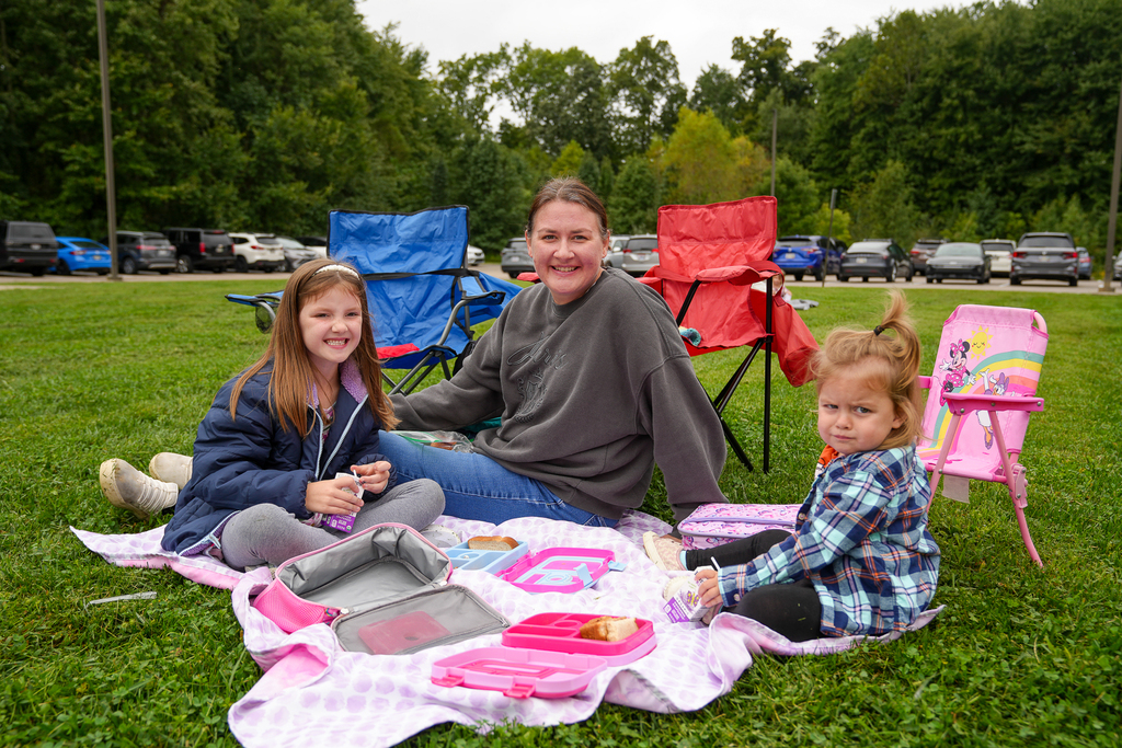 Two students and an adult smile while seated with their lunches on a blanket.