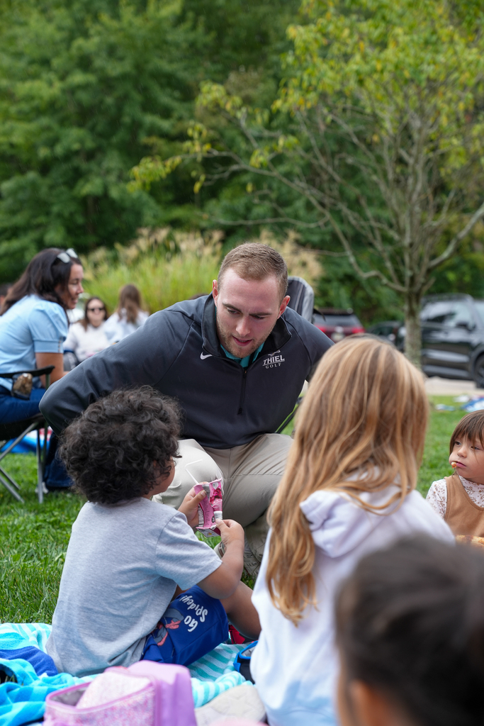 A staff member visits with a student and adults gathered on a picnic blanket.