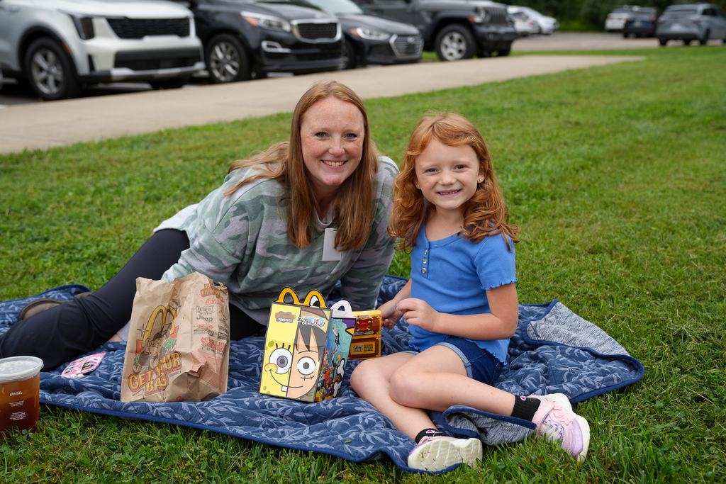 : An adult and student smile together while enjoying lunch on a blue blanket.