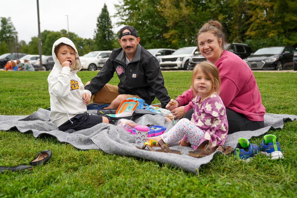 Two children and two adults enjoy a picnic lunch together on a gray blanket.