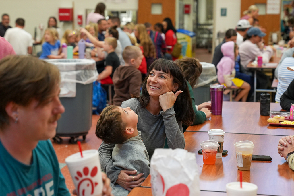 Families talk and eat breakfast together at tables in a busy school cafeteria.
