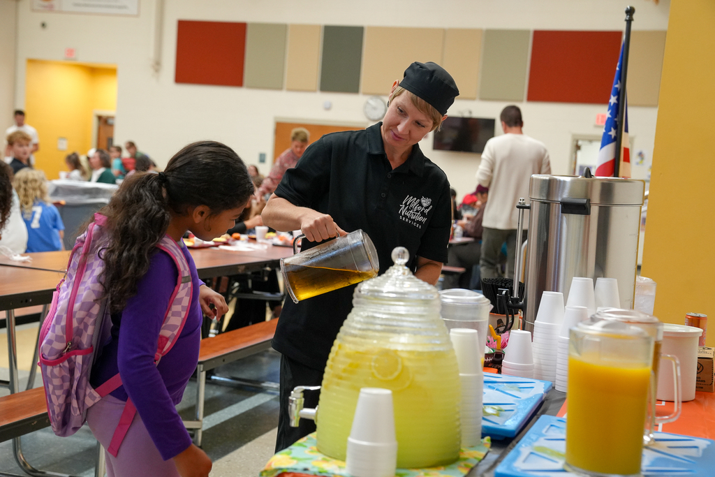A student fills a cup at a juice station while a staff member helps during breakfast.