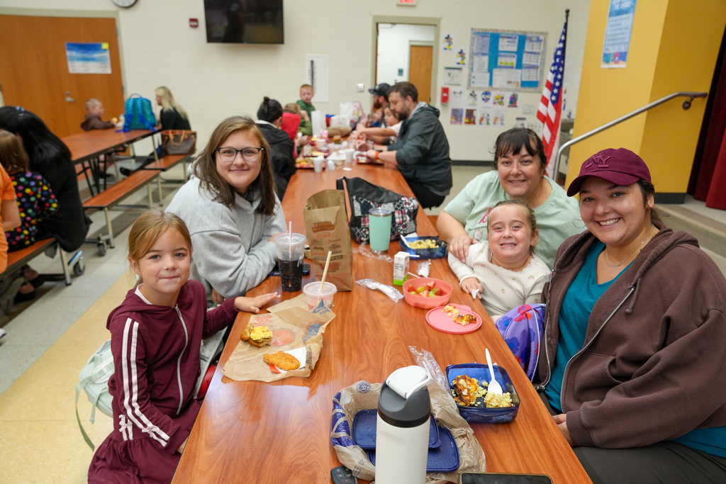Four family members smile at a cafeteria table while eating breakfast together.