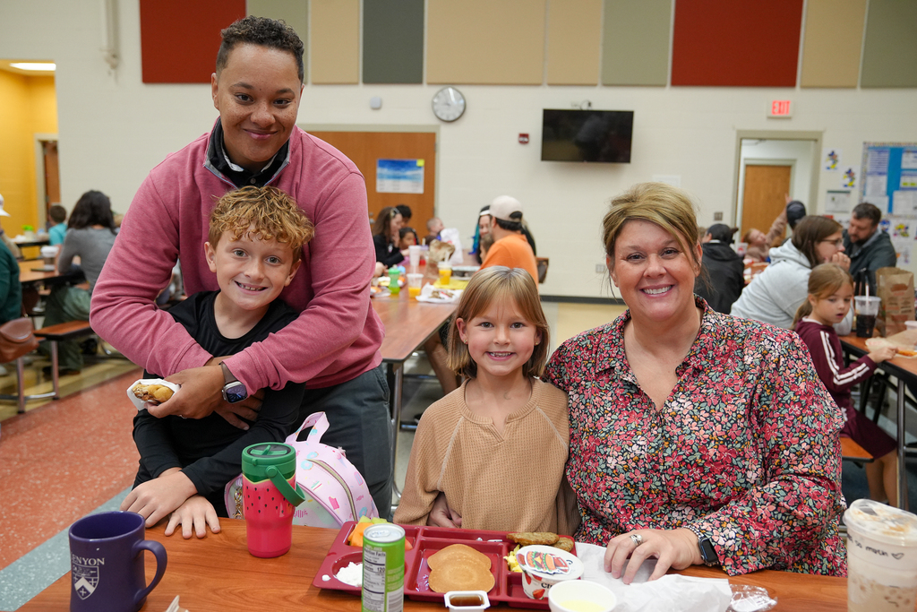 Two students and two adults smile around a cafeteria table filled with breakfast food.
