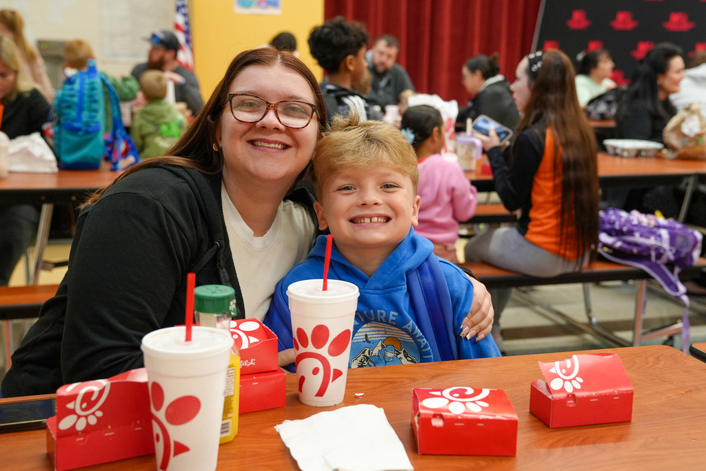 An adult and a student smile together at a cafeteria table with breakfast boxes and drinks.
