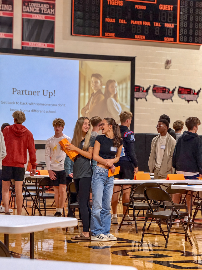 Students talk during a partner activity in the gym as a projected slide prompts them to meet someone from another school.