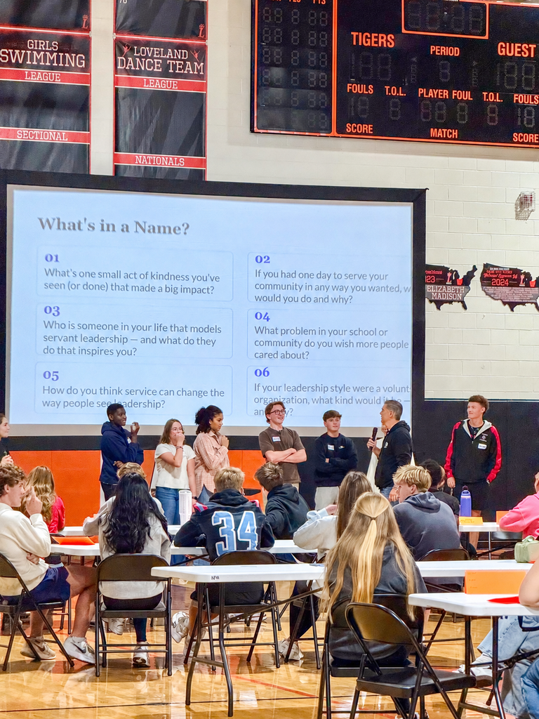 Students stand at the front of the gym for a leadership discussion while other participants watch from tables.