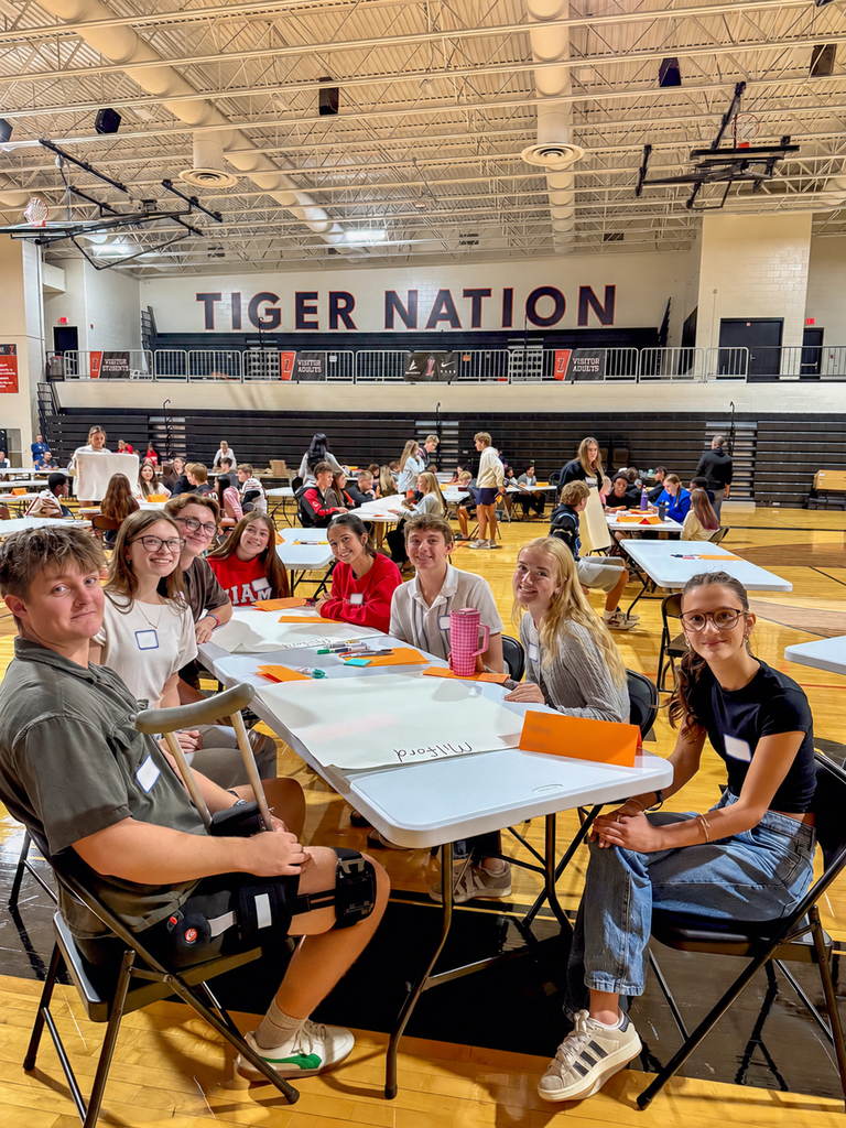 Milford students smile around a table with markers and activity materials as groups work throughout the gym.
