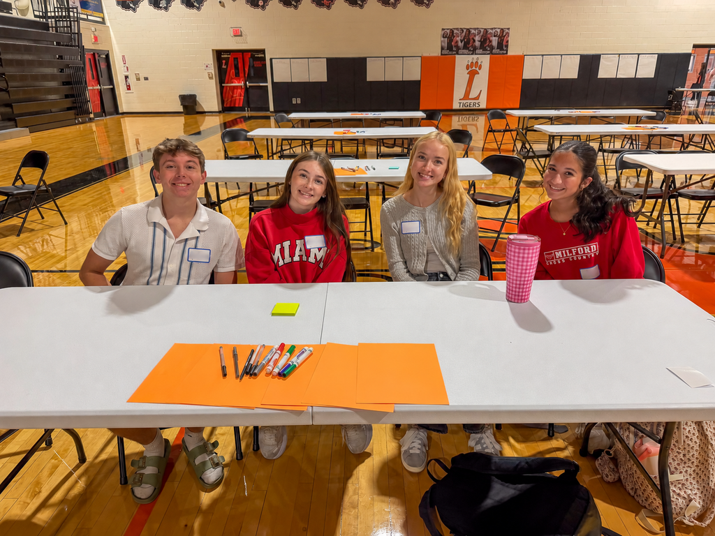 Milford students smile around a table with markers and activity materials as groups work throughout the gym.