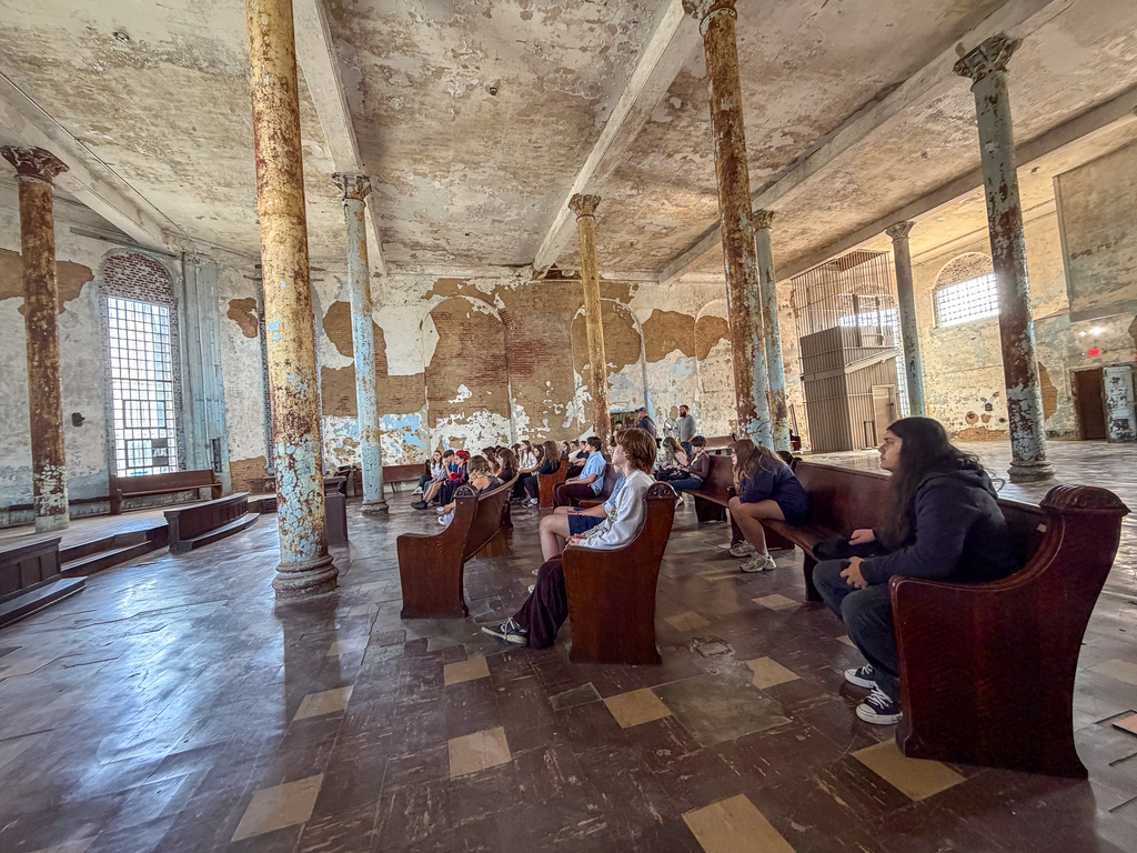 Students sit on wooden benches inside a large room at the reformatory, surrounded by tall columns and worn walls.