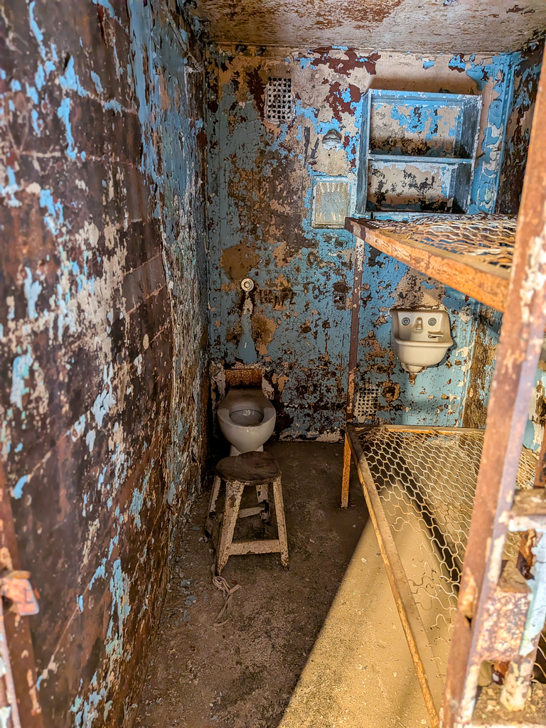 A former prison cell contains two metal bunks, a toilet and a sink beneath walls with peeling paint.