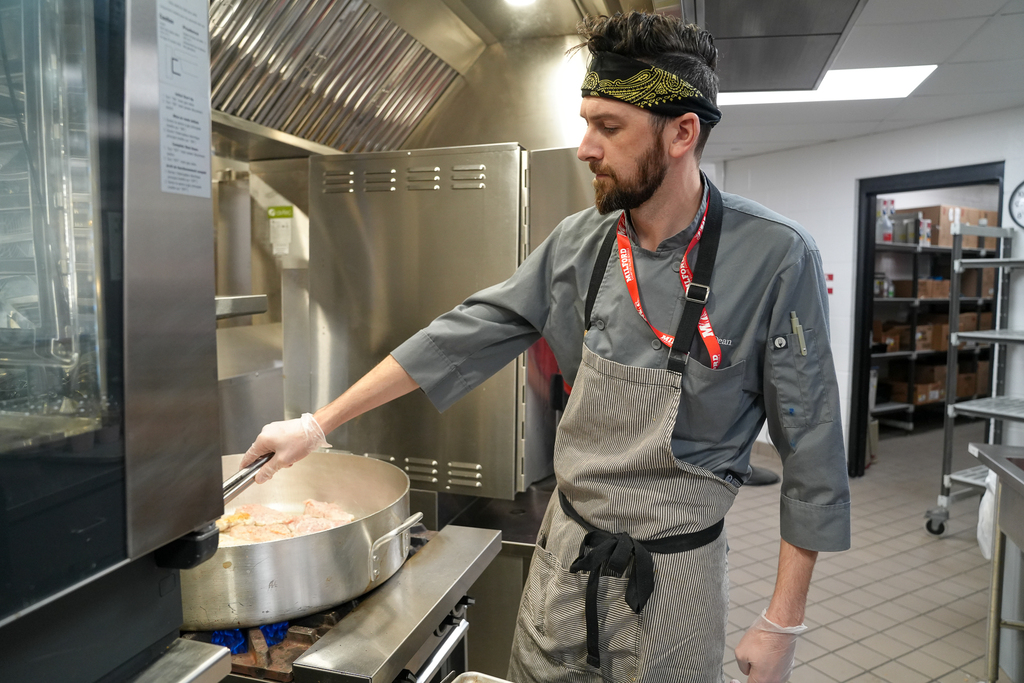 A Milford School Nutrition staff member cooks food in a large pan on the stove while preparing a meal in the school kitchen.