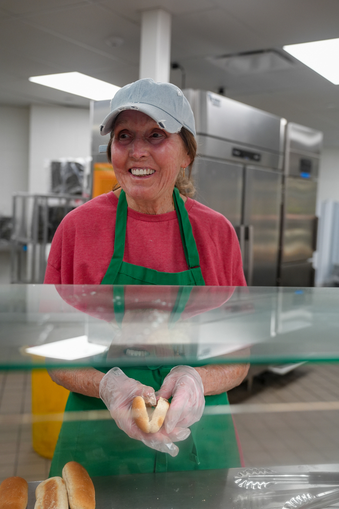 A Milford School Nutrition staff member smiles while preparing to serve a sandwich from behind the cafeteria counter.