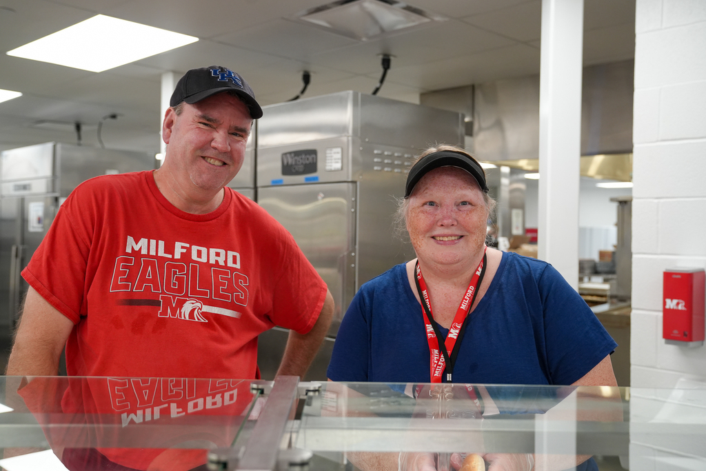 Two Milford School Nutrition staff members smile together behind the serving counter at Milford Middle School.
