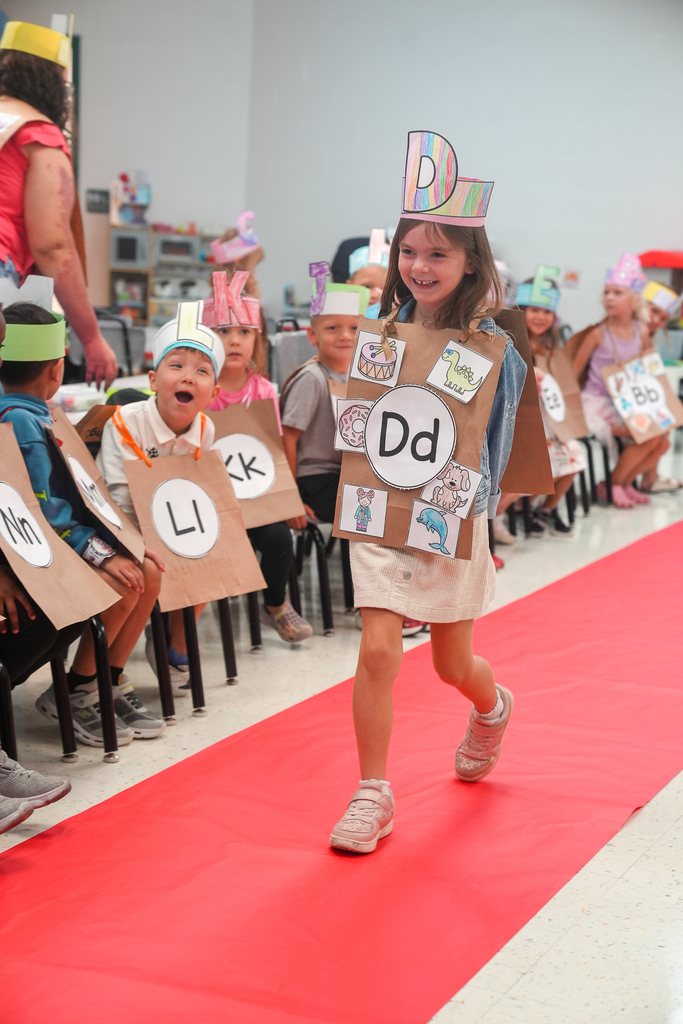 A kindergarten student walks down a red carpet in a handmade letter D costume as classmates watch during the ABC fashion show.