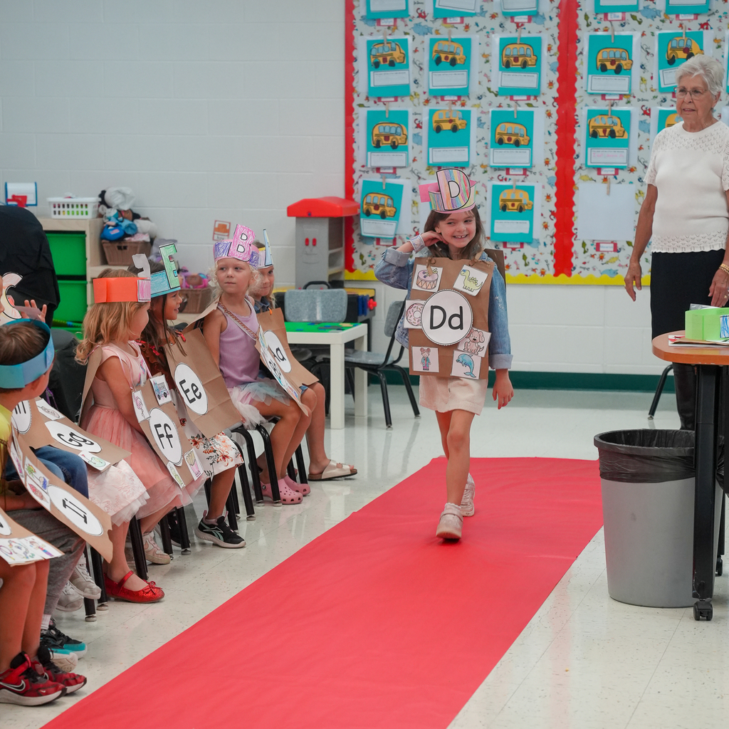 A kindergarten student smiles while walking the red carpet in a handmade letter D costume as classmates wearing other alphabet costumes watch.