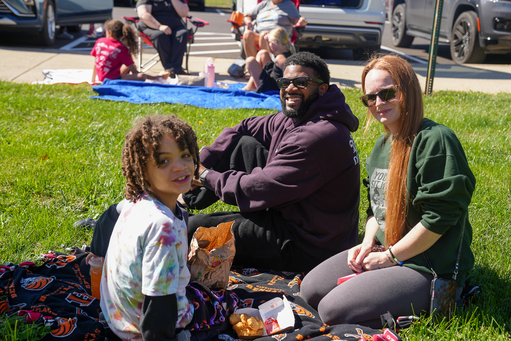 A student and two adults smile at the camera while sharing lunch on a Cincinnati Bengals blanket on the school lawn.