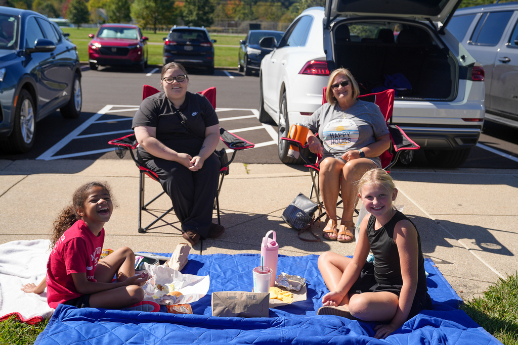 Two students smile while eating on a blue blanket, with two adults seated behind them in red folding chairs during Lunch on the Lawn.