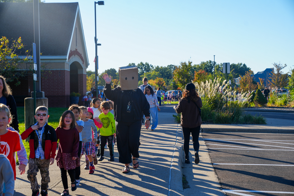 Students walk beside a staff member wearing a cardboard box with eyeholes over their head for “Secret Identity” spirit day. The staff member waves at the camera outside Mulberry Elementary.