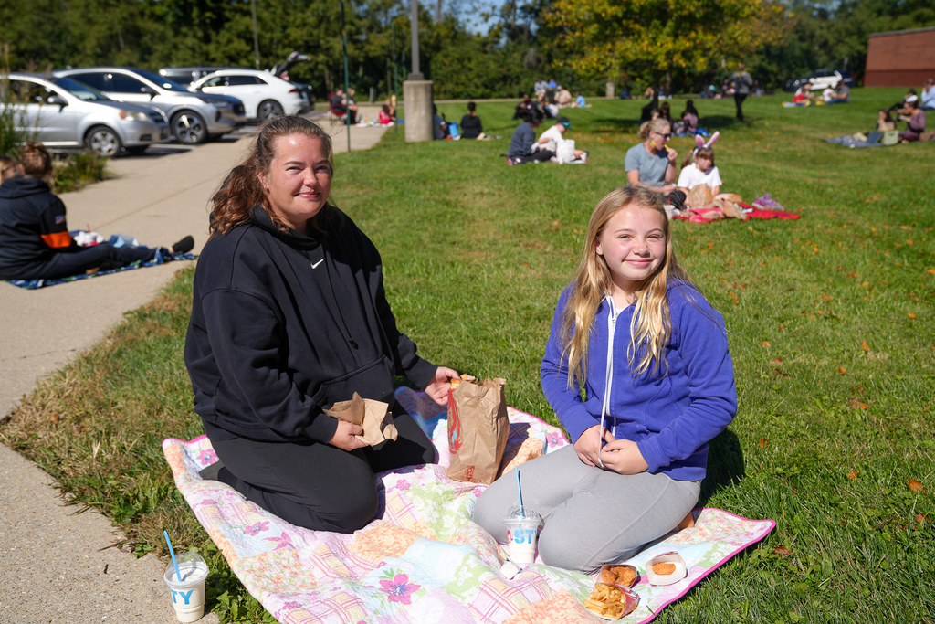 A student in a blue jacket and an adult in a black sweatshirt smile while sitting on a colorful quilt with their lunch. Other families picnic across the lawn behind them.