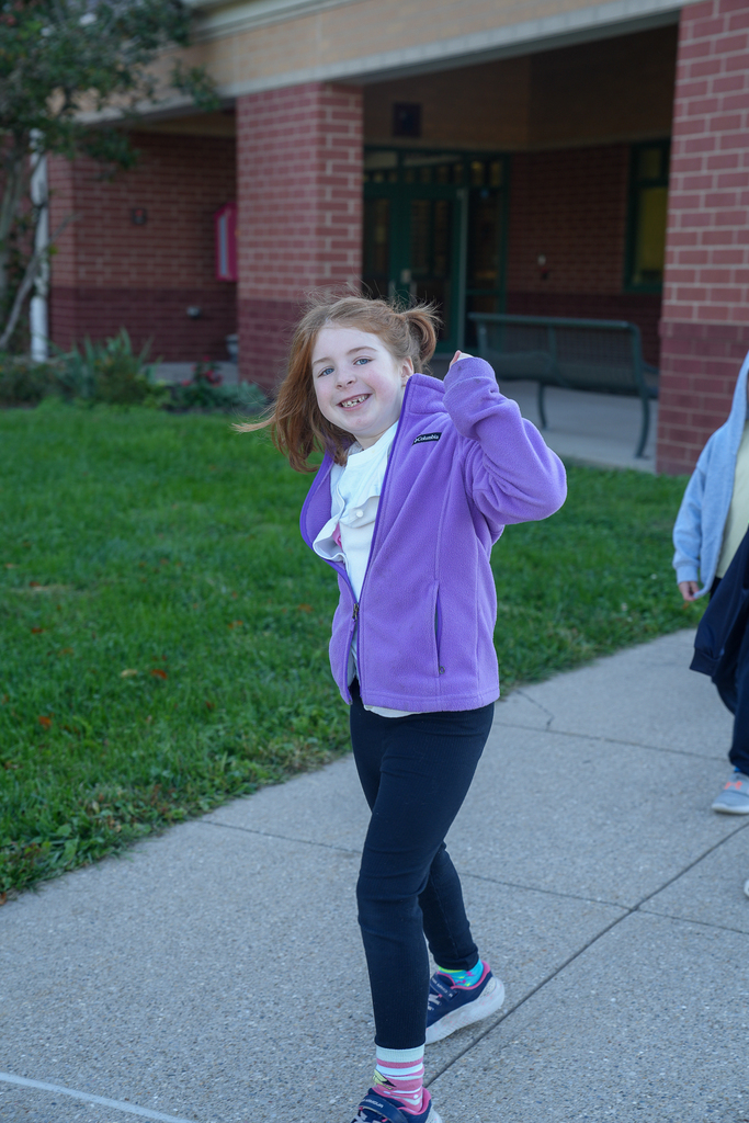 A student in a purple jacket smiles at the camera while walking along the sidewalk outside Mulberry Elementary during the Random Attendance Day reward.