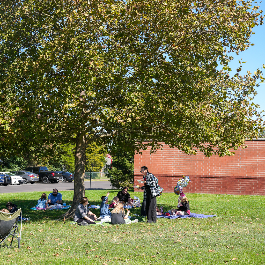 Students and adults gather on picnic blankets beneath a large shade tree outside Mulberry Elementary. A standing adult holds plates of food, and colorful balloons float beside one group.