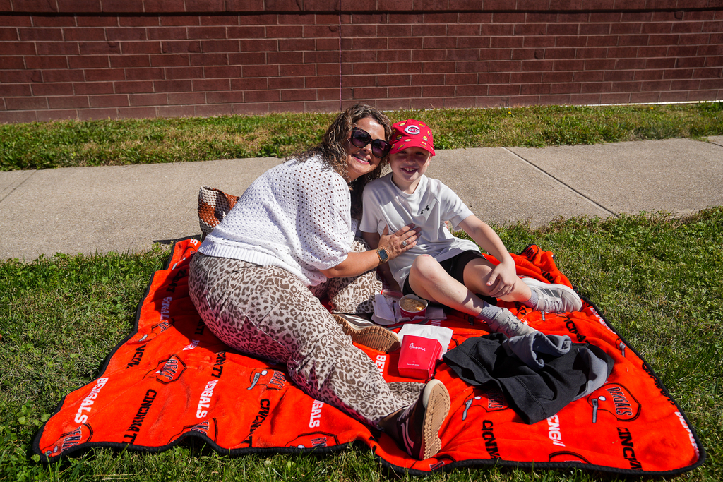 A student wearing a red Cincinnati Reds cap and an adult wearing sunglasses smile together on an orange Cincinnati Bengals blanket, with lunch beside them.
