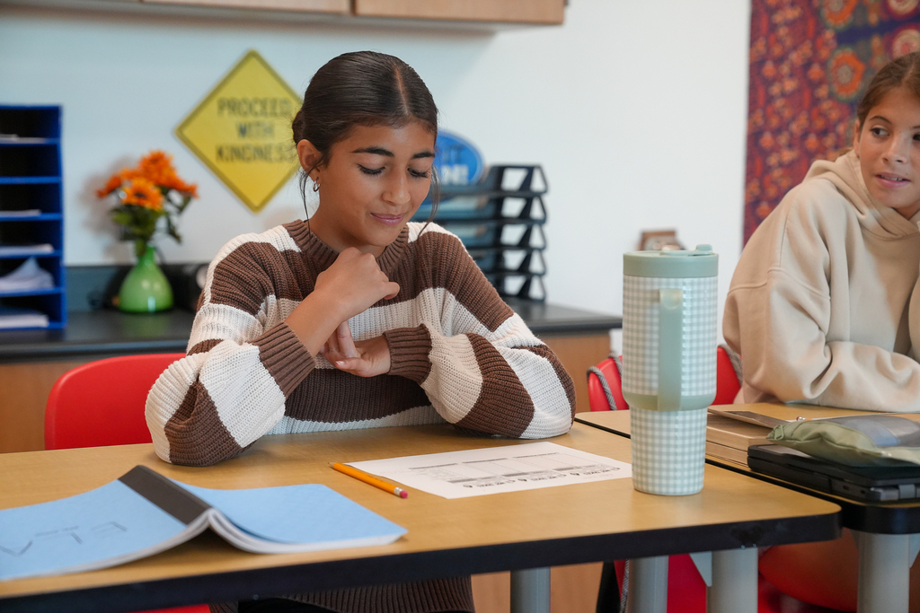 A student sits at a classroom table with her hands clasped during the Hot Takes activity, with papers and a pencil in front of her.
