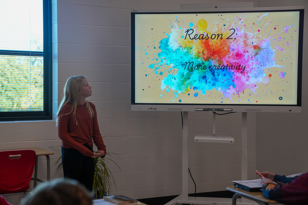 A student presents her argument beside a classroom screen displaying a colorful slide titled “Reason 2.”