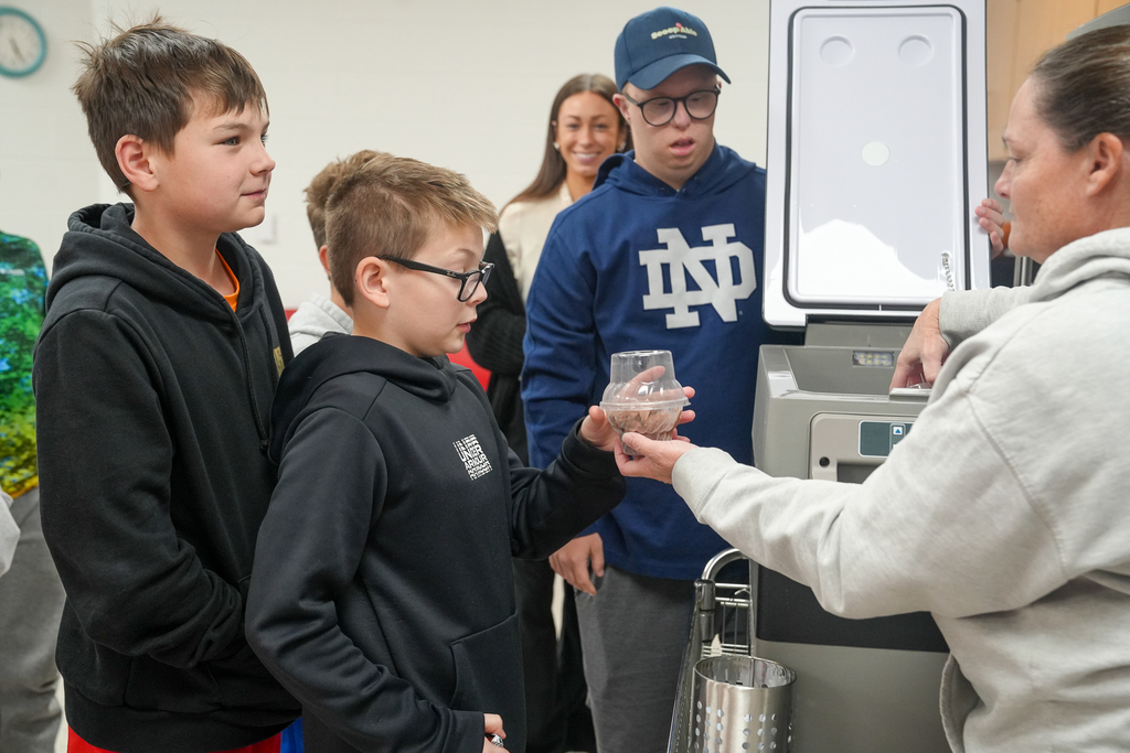 A ScoopAble representative hands a cup of ice cream to a Milford Middle School student as classmates gather nearby.