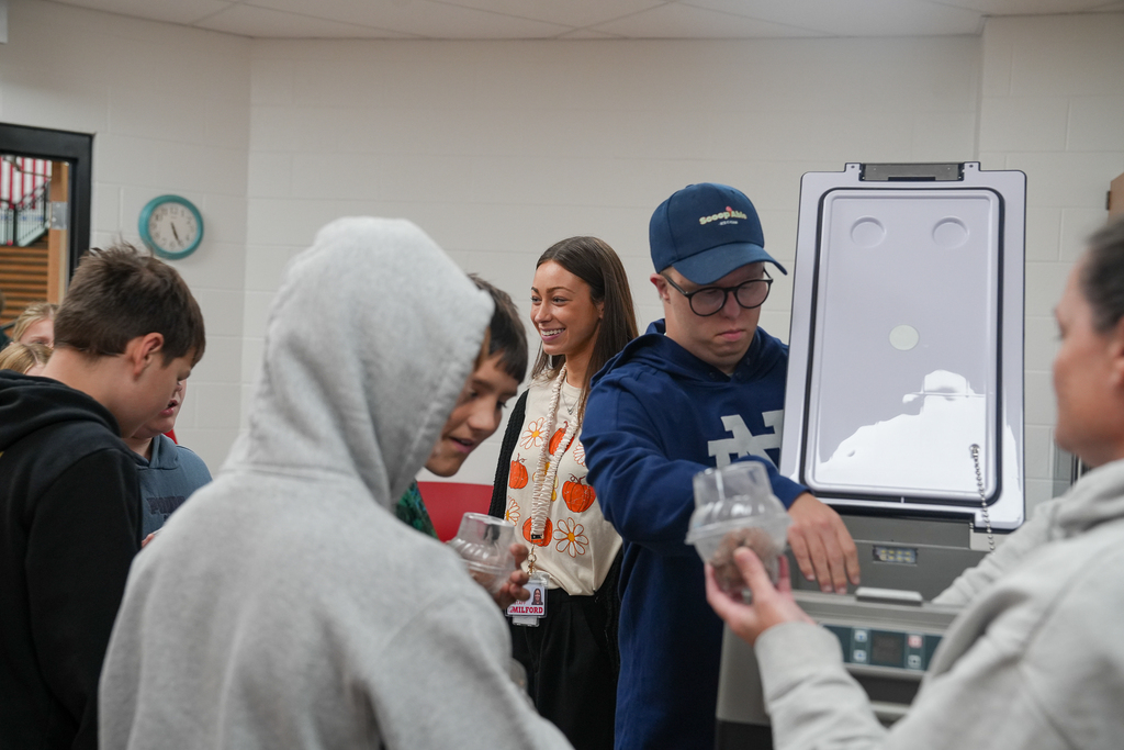 Milford Middle School students collect cups of ice cream from a ScoopAble representative beside an open cooler in their classroom.