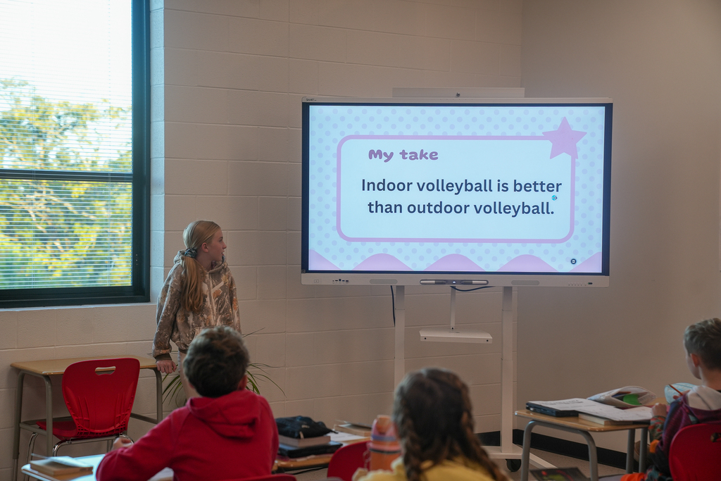 A student presents to classmates beside a slide reading, “My take: Indoor volleyball is better than outdoor volleyball.”