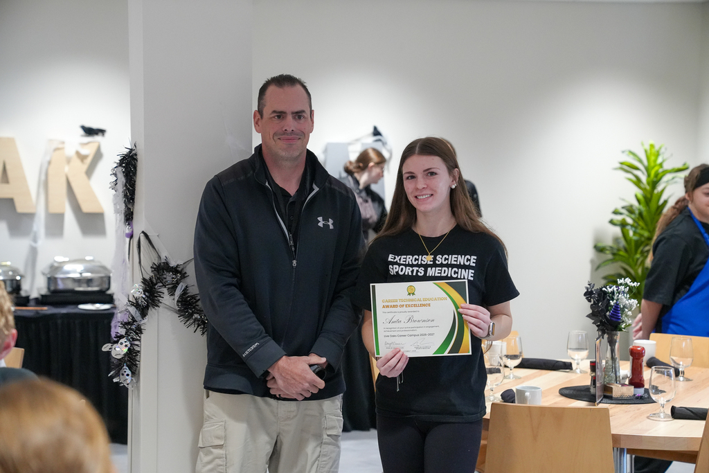 Milford student Anita Brownson smiles while holding her Career Technical Education Award of Excellence certificate alongside a Great Oaks staff member. Anita is wearing an Exercise Science and Sports Medicine shirt.