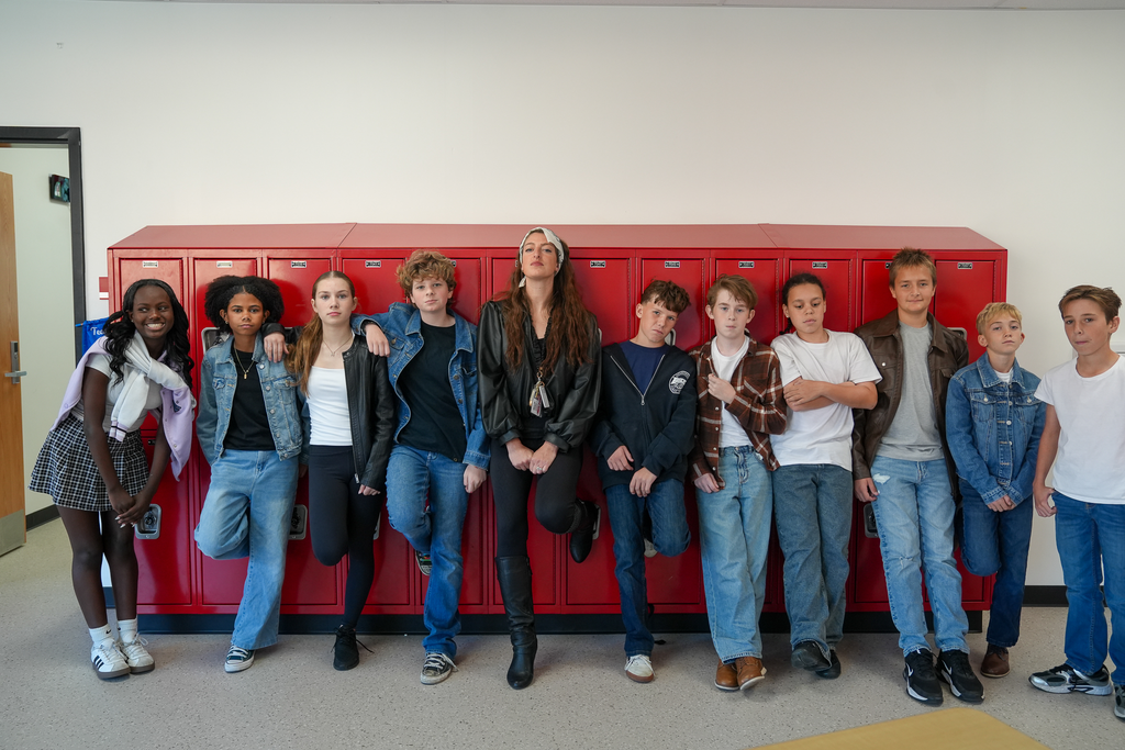 Students and a staff member wear outfits inspired by The Outsiders while posing along red lockers, several with one foot propped against the lockers.