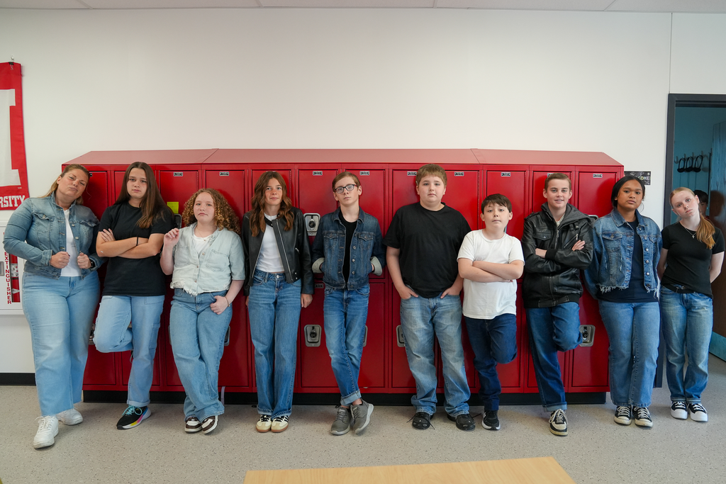 Students and a staff member wear outfits inspired by The Outsiders while posing along red lockers, several with one foot propped against the lockers.