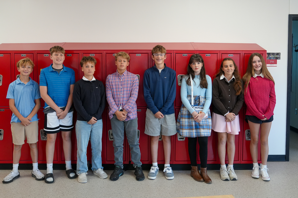 Eight students dressed as Socs pose in front of red lockers wearing polo shirts, sweaters, a plaid button-down shirt and skirts.