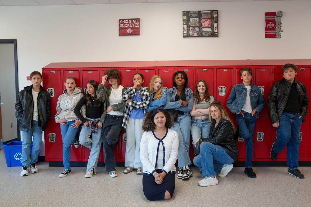 Students and a staff member wear outfits inspired by The Outsiders while posing along red lockers, several with one foot propped against the lockers.