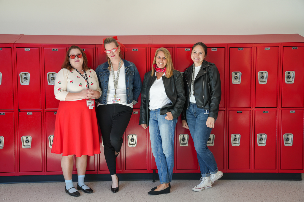 Four Milford Middle School staff members pose by red lockers wearing retro outfits, including a red skirt, cat-eye glasses, a denim jacket and black leather jackets.