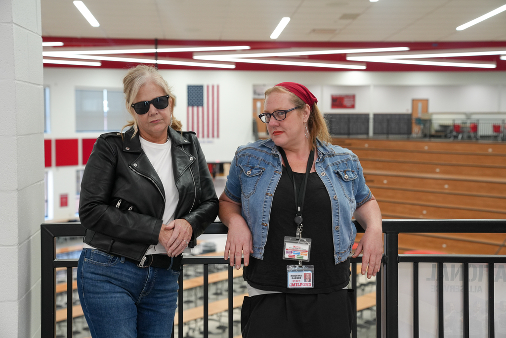 Two staff members lean against a railing in greaser-inspired outfits. One wears a black leather jacket and sunglasses; the other wears a denim vest and red headscarf.